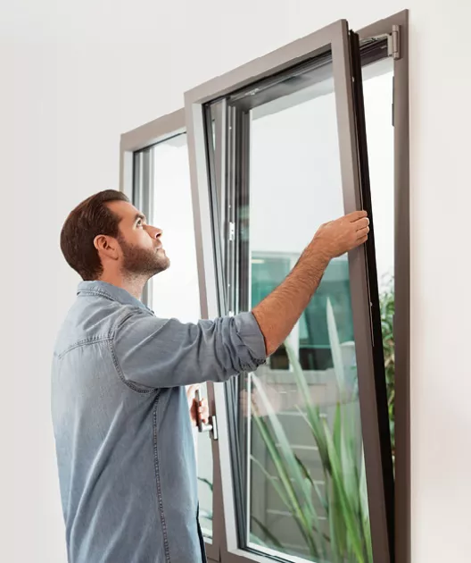 Homme inspectant une fenêtre partiellement ouverte, vêtu d'une chemise en jean, dans un environnement intérieur lumineux avec des plantes visibles à l'extérieur.