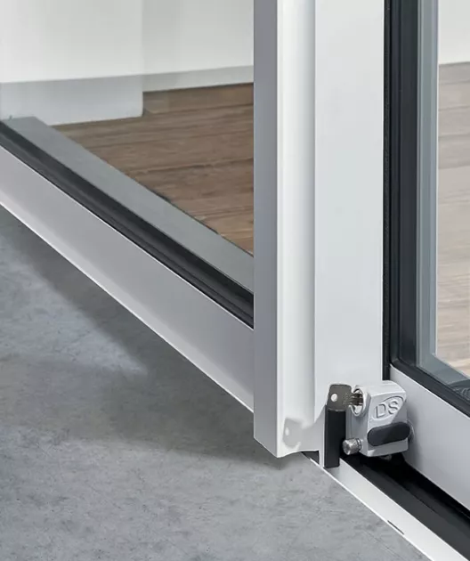 Fenêtre coulissante ouverte montrant le sol en bois intérieur, sol en béton extérieur, et un dispositif de verrouillage en métal gris au coin inférieur.