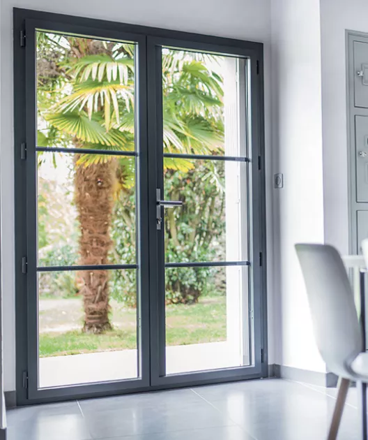 Porte-fenêtre vitrée ouverte sur un jardin verdoyant avec palmiers, ensoleillé. À l'intérieur, murs blancs et sol carrelé gris. Chaises modernes en bordure.
