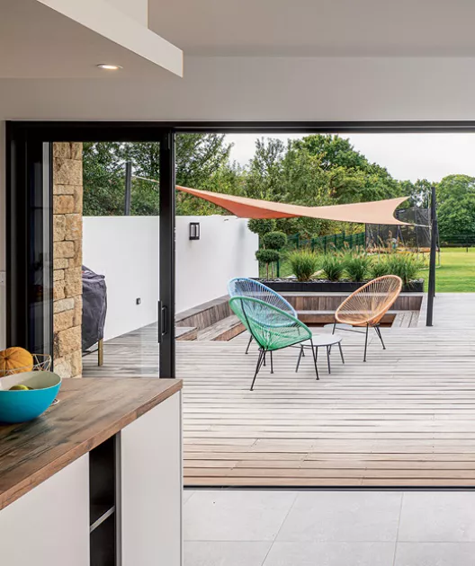 Deux chaises colorées sont posées sur une terrasse en bois, avec un auvent et un jardin verdoyant en arrière-plan.