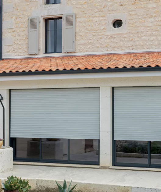 Les volets roulants baissés protègent de larges baies vitrées, sur la façade d'une maison en pierre avec un toit en tuiles.