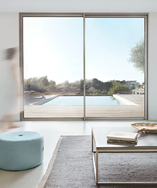 Grande baie vitrée encadrant une piscine extérieure, avec une personne floue marchant à gauche, dans un salon élégant doté de meubles modernes.