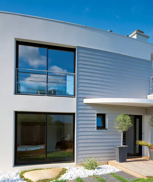 Maison moderne avec grandes fenêtres offrant vue sur jardin, façade grise et blanche, arborant des plantes décoratives près de l'entrée.