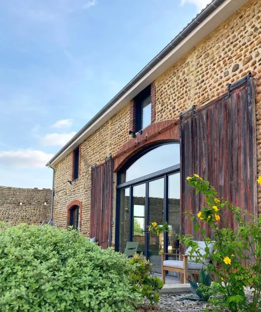 Grande bâtisse en pierre rénovée avec baies vitrées et portes en bois, entourée de verdure sous un ciel bleu.