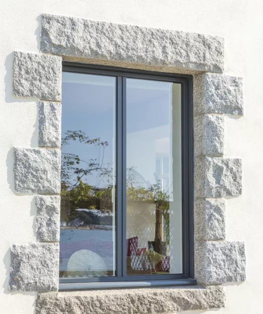 Fenêtre à double vitrage, entourée de pierres de taille grises, reflétant un jardin extérieur avec arbres et quelques objets intérieurs visibles derrière le verre.