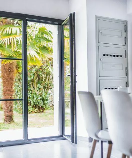 Porte-fenêtre ouverte laissant entrer la lumière naturelle dans une pièce moderne, révélant un jardin avec des palmiers en arrière-plan. Des chaises blanches sont en premier plan.