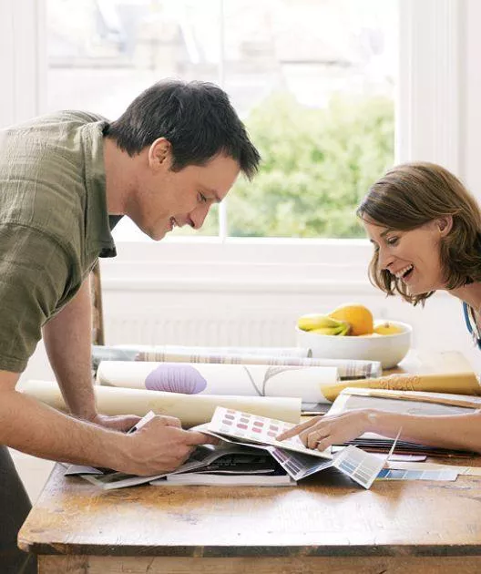 Deux personnes examinent des échantillons de papier peint et de couleurs sur une table, souriant et discutant, dans un salon ensoleillé avec un bol de fruits en arrière-plan.
