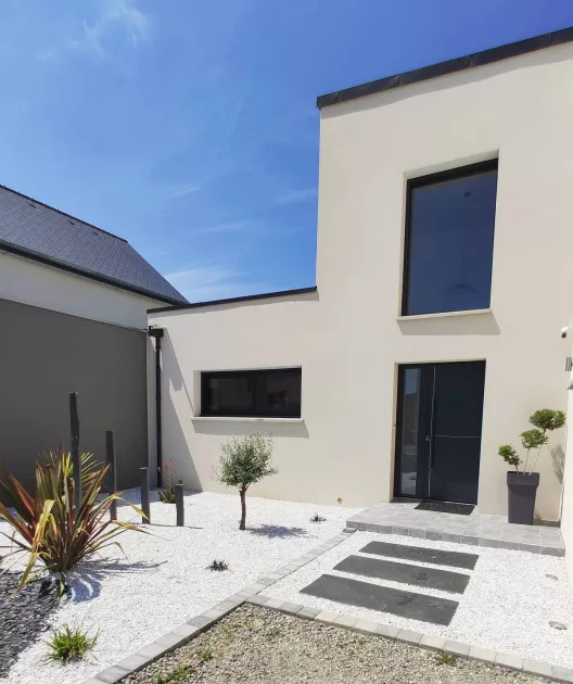 Maison moderne en façade crème, large fenêtre, porte noire, petit jardin de graviers blancs, plantes décoratives, allée avec dalles grises, ciel bleu.