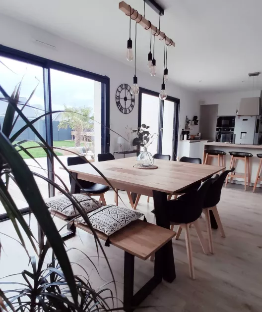 Table à manger en bois entourée de chaises noires, surmontée de suspensions lumineuses, située dans une salle à manger lumineuse avec vues sur jardin par grandes baies vitrées.