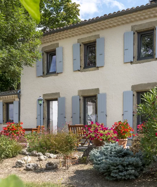 Maison blanche à volets gris entourée de fleurs colorées et de buissons, située dans un jardin ensoleillé avec un banc en bois devant.