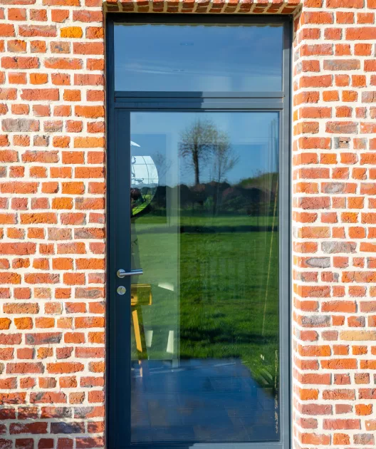 Porte vitrée reflétant un arbre dans un paysage verdoyant, encadrée par un mur en briques rouges.