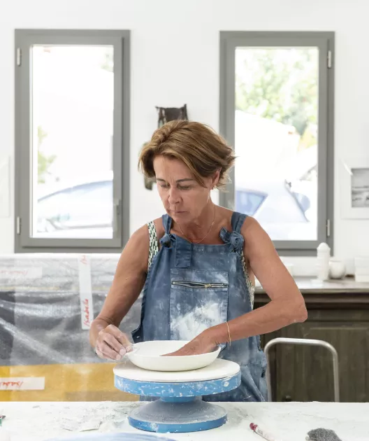 Une femme crée une céramique sur un plateau tournant bleu dans un atelier avec des fenêtres en arrière-plan.