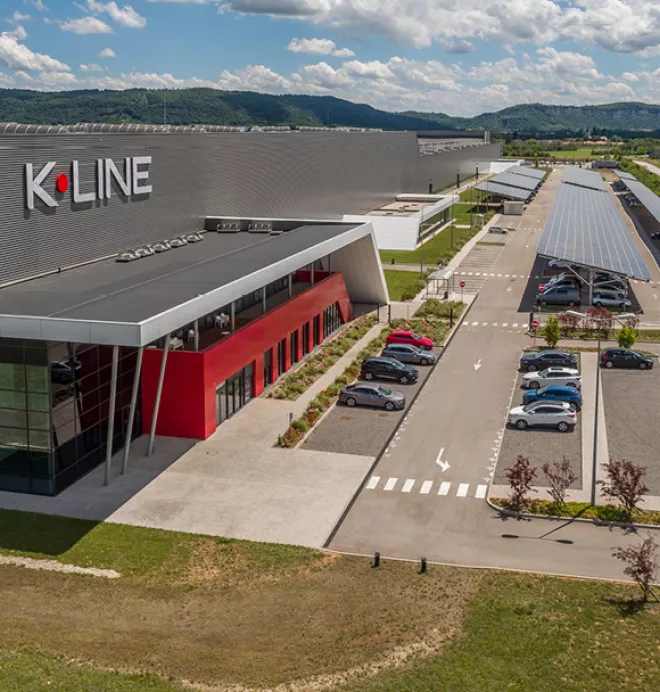 Un bâtiment moderne avec un panneau "K.LINE", des voitures garées, entouré d'un paysage verdoyant et montagneux sous un ciel bleu parsemé de nuages.