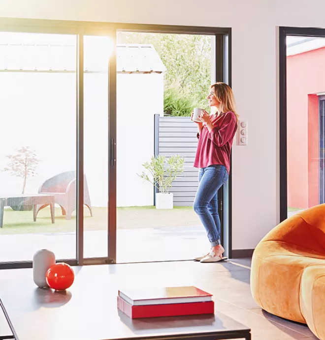 Une femme, tenant une tasse, se tient près d'une porte vitrée ouverte, regardant dehors dans un jardin ensoleillé, depuis un salon moderne.