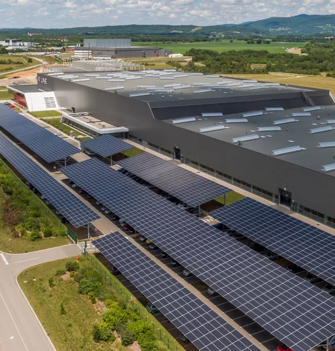 Un vaste bâtiment industriel avec des panneaux solaires en toiture et un parking couvert de structures de panneaux solaires, situé en pleine campagne verdoyante et vallonnée.