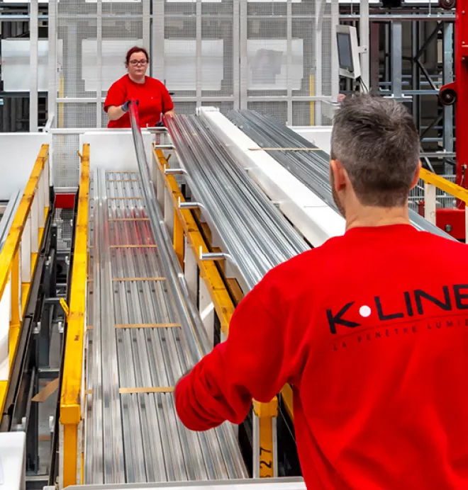 Deux ouvriers en tenues rouges manipulent de longues barres métalliques sur une chaîne de production dans une usine moderne.