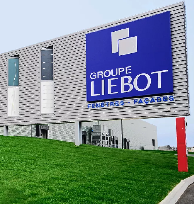 Un grand panneau bleu du Groupe Liebot surplombe une pelouse verte, indiquant une entreprise spécialisée dans les fenêtres et façades, devant un bâtiment industriel moderne.