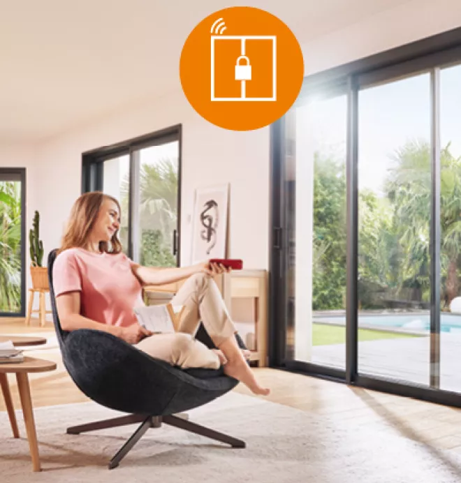 Une femme assise dans un fauteuil utilise une télécommande pour ouvrir une baie vitrée, donnant sur un jardin ensoleillé avec piscine.