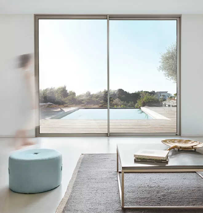 Grande baie vitrée encadrant une piscine extérieure, avec une personne floue marchant à gauche, dans un salon élégant doté de meubles modernes.