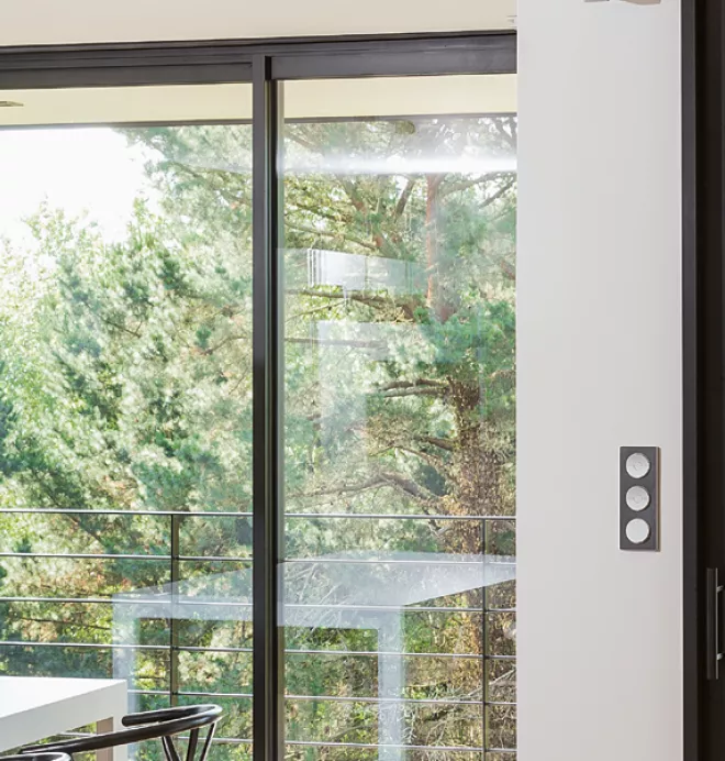 Des grandes baies vitrées laissent entrer la lumière dans une pièce moderne, bordée d'arbres visibles à l'extérieur. Deux suspensions noires pendent au-dessus d'une table blanche.
