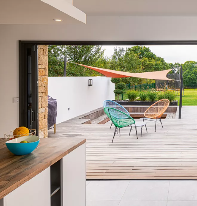 Chaises colorées disposées sur une terrasse extérieure en bois, abritées par une voile d'ombrage, donnent sur un jardin verdoyant.