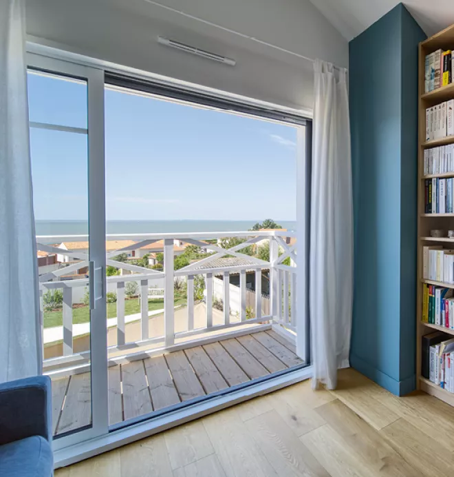Une grande baie à galandage vitrée ouverte donnant sur un balcon en bois, avec vue sur la mer et des toits de maisons proches, intégrée à un salon lumineux avec des étagères remplies de livres.