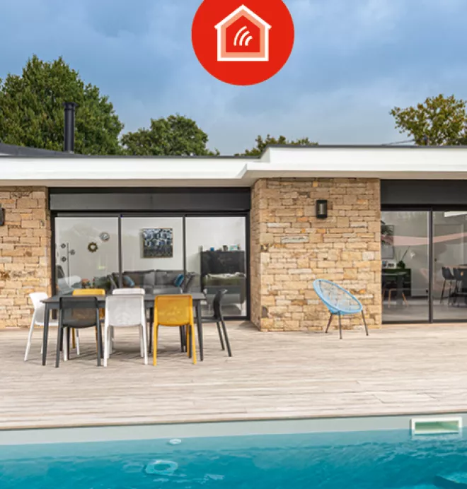 Piscine devant une maison moderne en pierres, flanquée de chaises colorées et d’une table sur une terrasse en bois, sous un ciel nuageux.