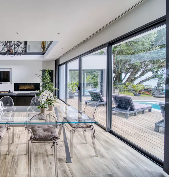 Table en verre, entourée de chaises transparentes, placée dans un salon moderne avec vue sur une terrasse en bois et une piscine extérieure.