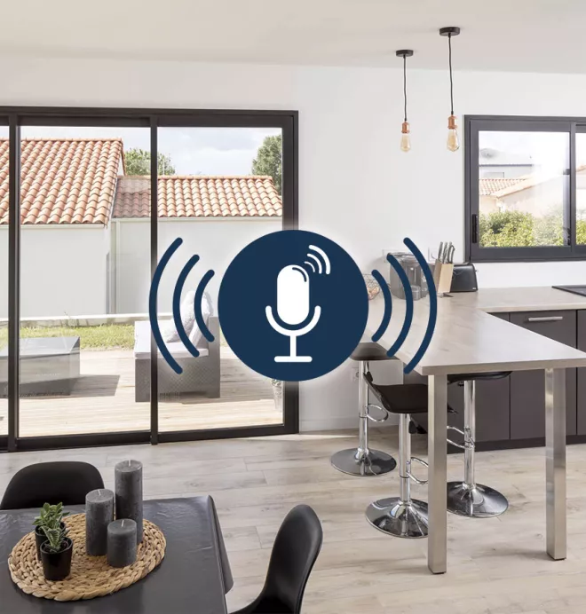 Une cuisine moderne avec îlot et tabourets; deux suspensions lumineuses; baie vitrée ouverte sur une terrasse avec mobilier extérieur; icône de microphone indiquant une fonctionnalité audio.