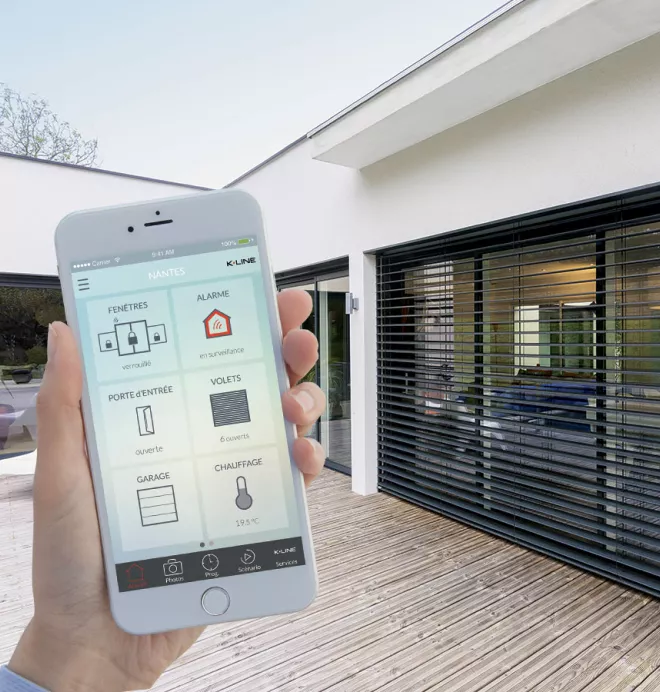 Un smartphone contrôle les dispositifs de sécurité et de confort d’une maison moderne avec toit-terrasse par une application de domotique.