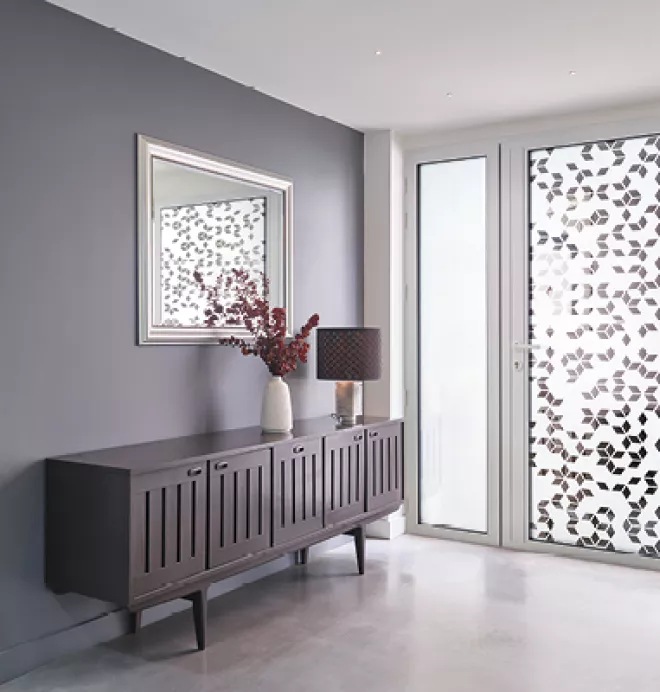 Buffet en bois avec vase et lampe, devant une fenêtre décorée de motifs floraux. Miroir encadré au-dessus, murs gris et sol clair. Atmosphère moderne et lumineuse.