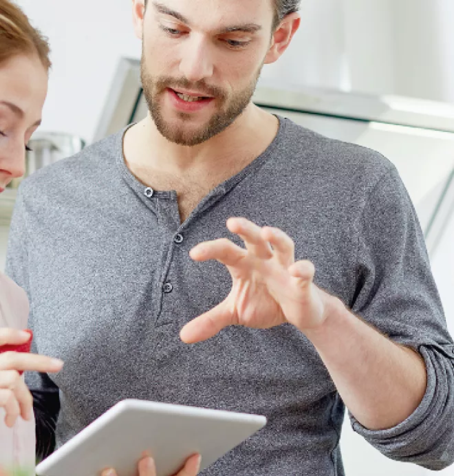 Deux personnes consultent une tablette dans une cuisine moderne, échangeant des informations et discutant activement.