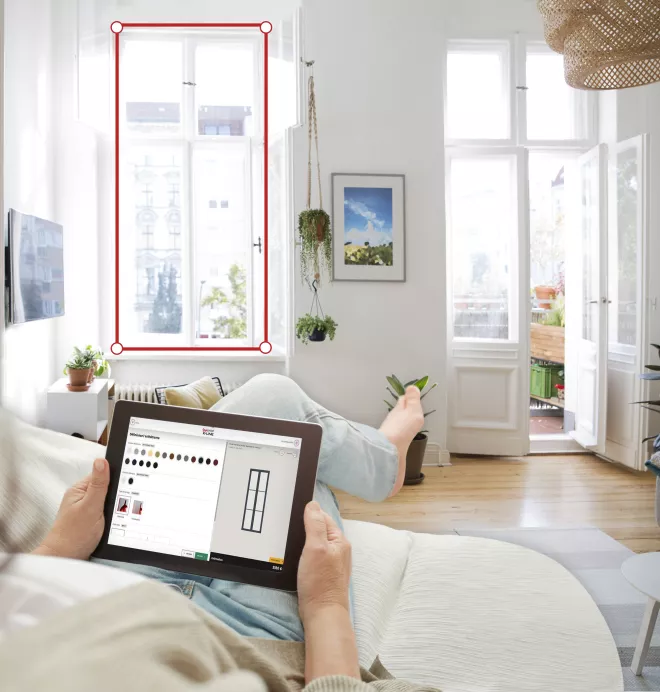 Tablette tenue par une personne allongée, consultant une application de personnalisation, dans un salon lumineux avec de grandes fenêtres ouvertes sur l'extérieur.