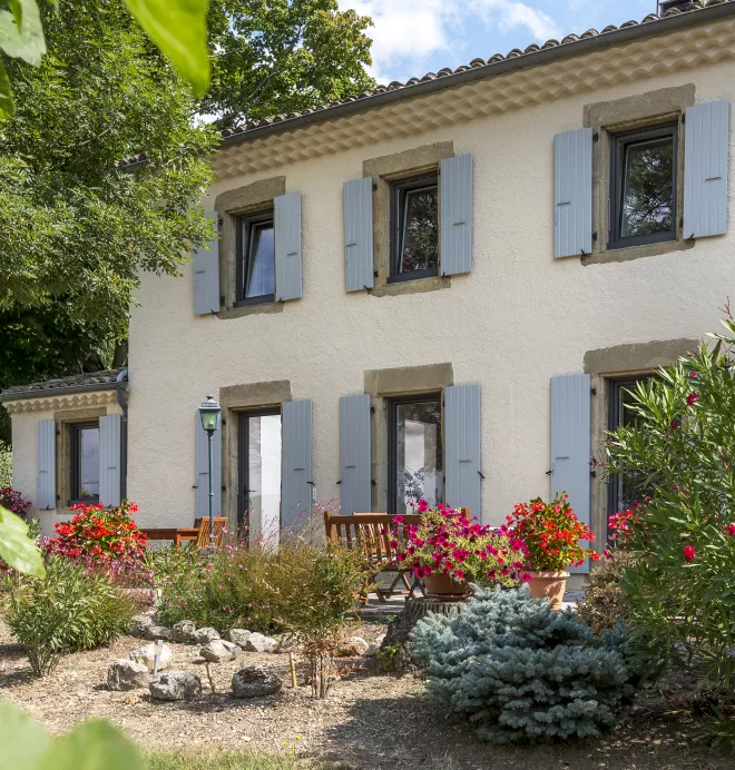 Maison blanche à volets gris entourée de fleurs colorées et de buissons, située dans un jardin ensoleillé avec un banc en bois devant.