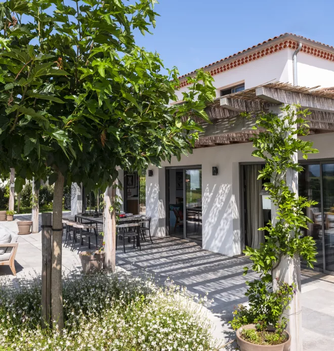 Maison avec terrasse ombragée, entourée de plantes vertes et fleurs blanches, située sous un ciel bleu clair. Tables et chaises en métal sont disposées pour des repas en plein air.