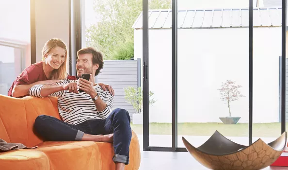 Un homme et une femme sourient et regardent un smartphone, assis sur un canapé orange dans un salon lumineux avec grande baie vitrée.