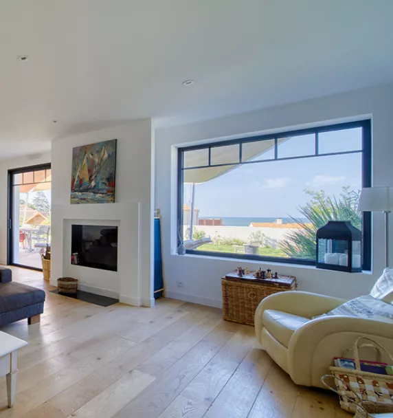 Salon moderne meublé avec un canapé gris, un fauteuil beige, et une cheminée, offrant une vue sur la mer par une grande baie vitrée.