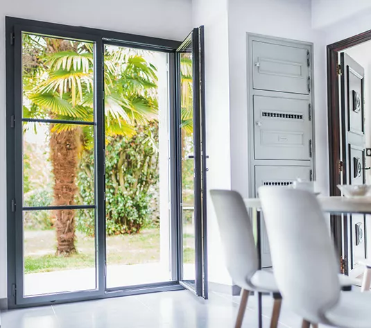 Une porte vitrée s'ouvre sur un jardin luxuriant avec des palmiers, laissant entrer beaucoup de lumière dans une salle à manger moderne.