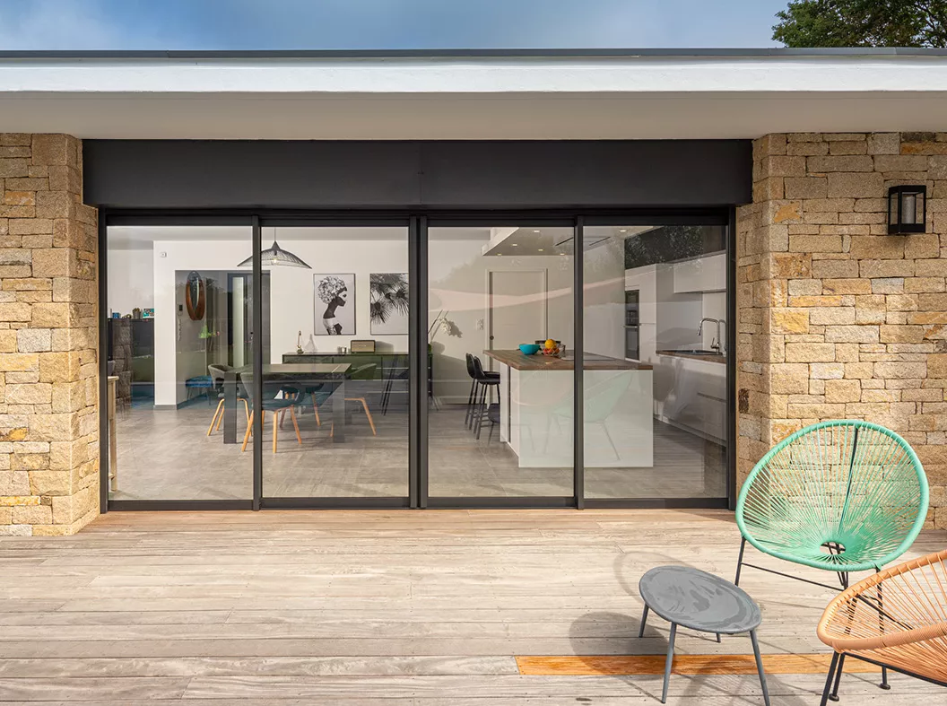 Une terrasse en bois avec des fauteuils colorés jouxte une maison moderne aux grandes baies vitrées, dévoilant une cuisine et une salle à manger intérieur élégantes et lumineuses.