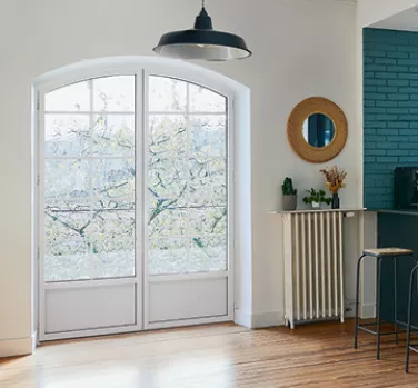 Porte-fenêtre vitrée donnant sur jardin, laissant pénétrer la lumière dans un salon avec parquet, tabouret haut, mur turquoise et décoration minimaliste, dont miroir rond et fleurs séchées.