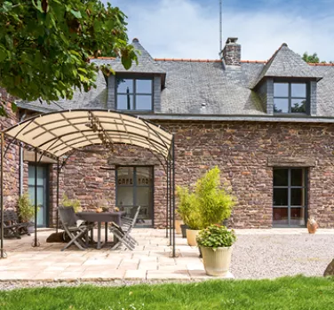 Une maison en pierre avec des fenêtres mansardées, un espace extérieur aménagé sous une pergola, situé dans un jardin verdoyant.