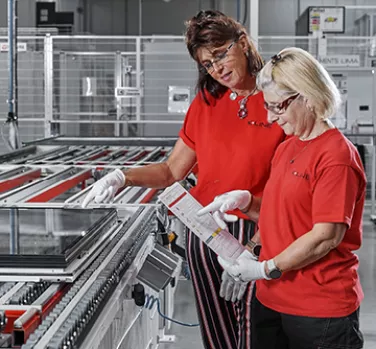 Deux personnes en uniforme rouge inspectent un document devant une chaîne de montage industrielle, portant des gants blancs dans un environnement d'usine.