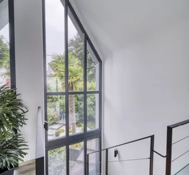 Grande fenêtre vitrée laisse entrer la lumière naturelle, illuminant un escalier moderne avec des plantes et une vue sur un jardin verdoyant.