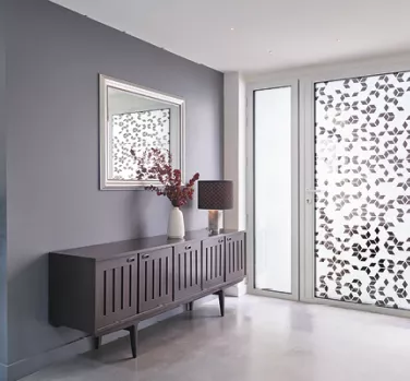 Buffet en bois avec vase et lampe, devant une fenêtre décorée de motifs floraux. Miroir encadré au-dessus, murs gris et sol clair. Atmosphère moderne et lumineuse.