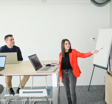 Une femme en blazer orange, debout près d'un tableau blanc, présente des informations à deux personnes assises avec des ordinateurs portables dans une salle de réunion.