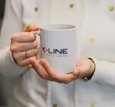 Une main tient une tasse blanche marquée "K.LINE" dans un environnement intérieur, avec un vêtement blanc décoré de petits cœurs dorés en arrière-plan.