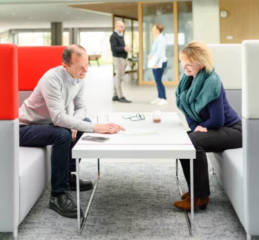 Deux personnes discutent autour d'une table basse dans un espace de bureau lumineux, tandis que d'autres travaillent en arrière-plan.