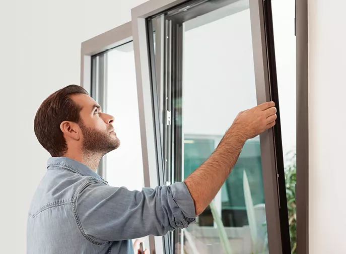 Homme ouvre une fenêtre basculante dans une pièce lumineuse, vêtu d'une chemise en jean bleu.