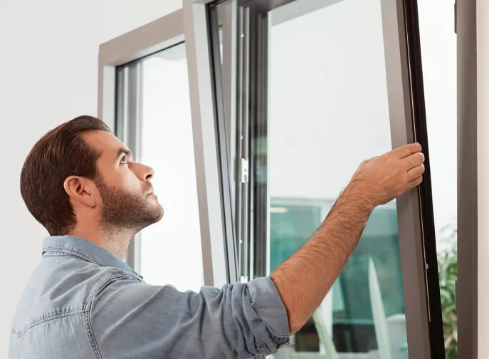 Un homme ouvre une fenêtre moderne dans une pièce lumineuse, révélant un environnement extérieur avec des plantes visibles en arrière-plan.
