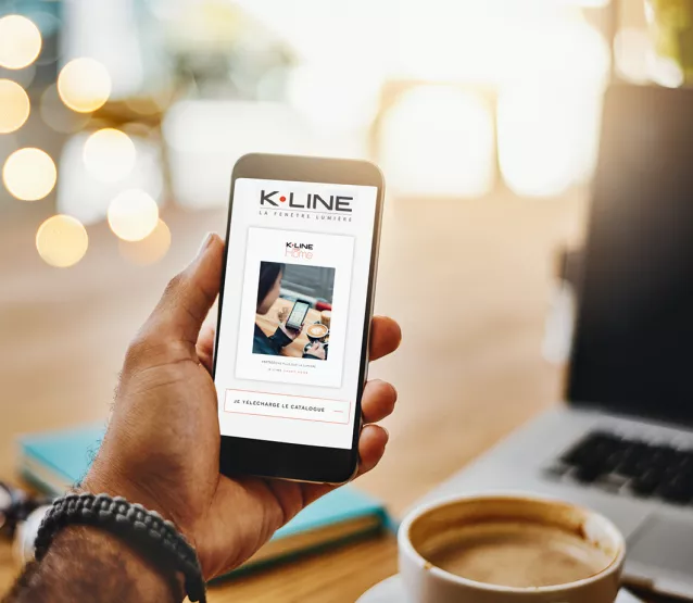 Main tenant un smartphone avec une publicité pour K-LINE, assis à une table avec un ordinateur portable et une tasse de café à côté.